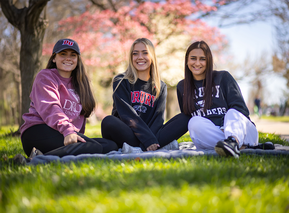 Three female students looking at the camera, smiling. They are sitting in the grass.